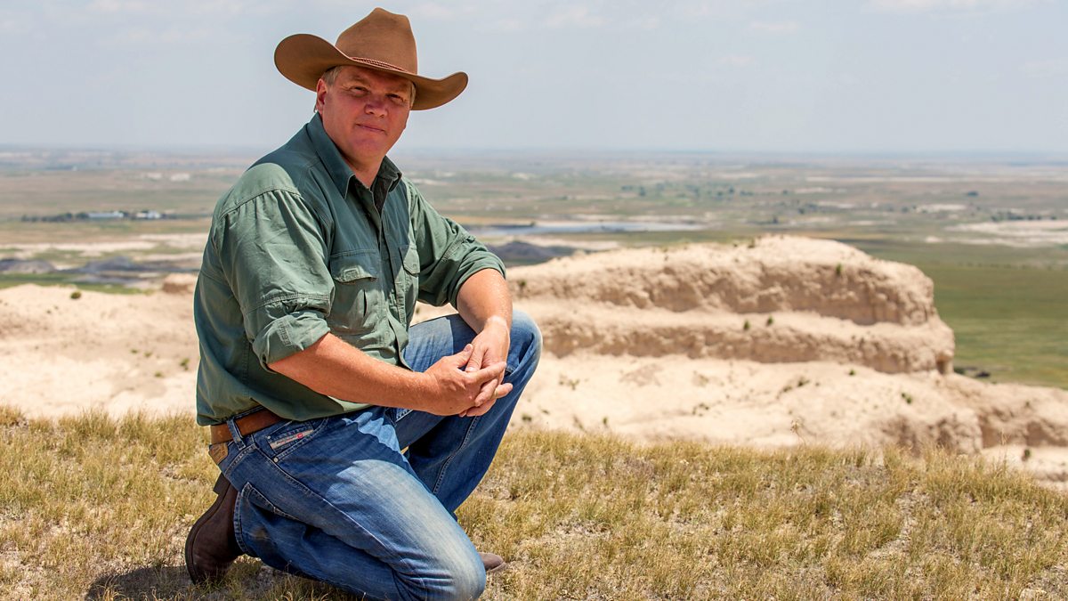 Man in a cowboy hat and green shirt sitting on a grassy hill with a scenic landscape in the background